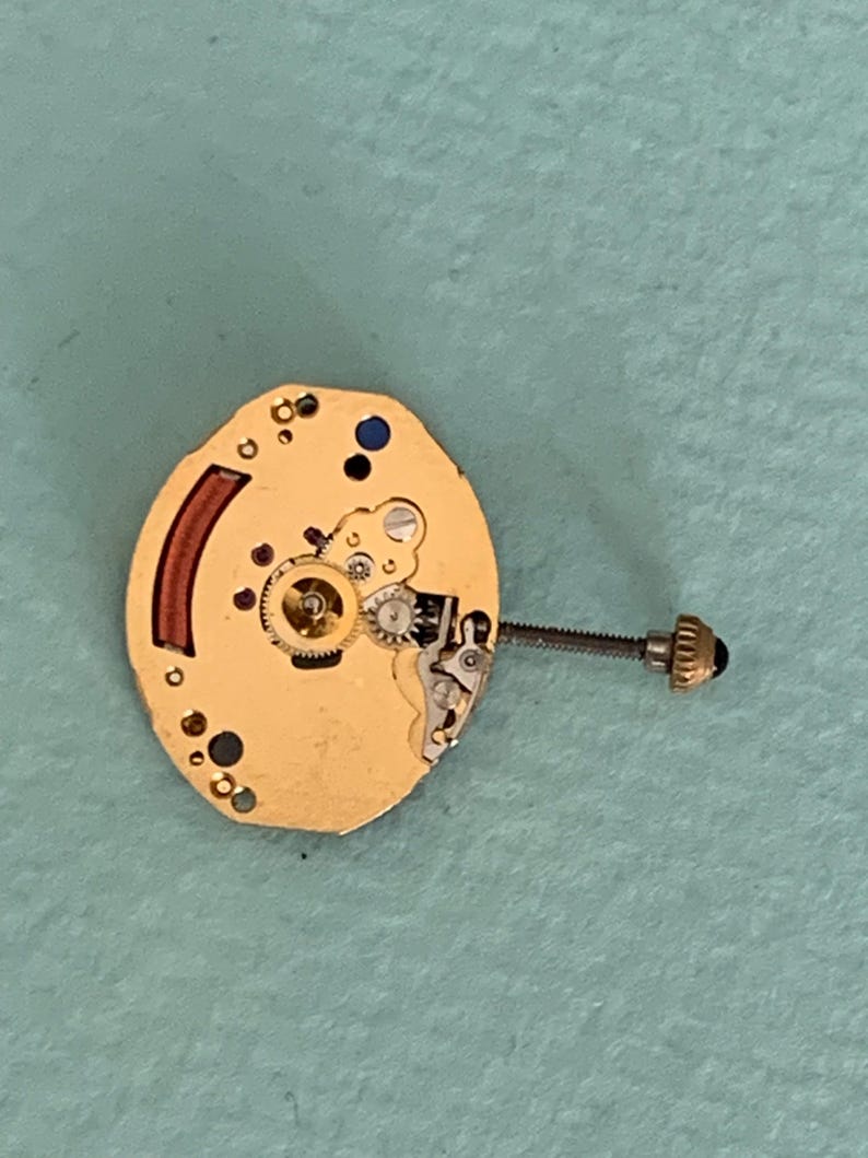 May include: A gold-colored watch movement with intricate gears, a red curved component, and a screw with a black head. The watch mechanism is oval-shaped and has small holes and blue accents. The image is taken from a top-down perspective.