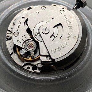 Pu&ograve; includere: Primo piano di un movimento di orologio argentato con la scritta "ORIENT 21J 46941" e "JAPAN". Il meccanismo comprende ingranaggi, viti e un albero di carica. Il movimento dell'orologio &egrave; in un contenitore trasparente e circolare.