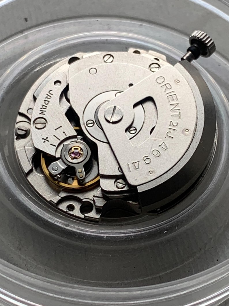 Pu&ograve; includere: Primo piano di un movimento di orologio argentato con la scritta "ORIENT 21J 46941" e "JAPAN". Il meccanismo comprende ingranaggi, viti e un albero di carica. Il movimento dell'orologio &egrave; in un contenitore trasparente e circolare.