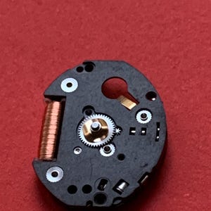 May include: Close-up of a watch movement component. The black circular piece features a copper coil, small gears, and various silver and gold-colored components. The background is a solid red.