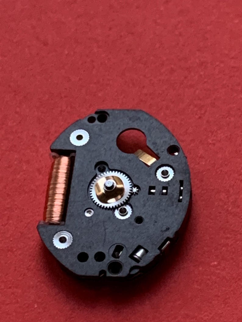 May include: Close-up of a watch movement component. The black circular piece features a copper coil, small gears, and various silver and gold-colored components. The background is a solid red.
