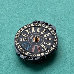 May include: A black watch calendar wheel with gold-colored text and numbers. The wheel displays the days of the week in multiple languages and the dates from 1 to 31. The wheel is set against a teal background.
