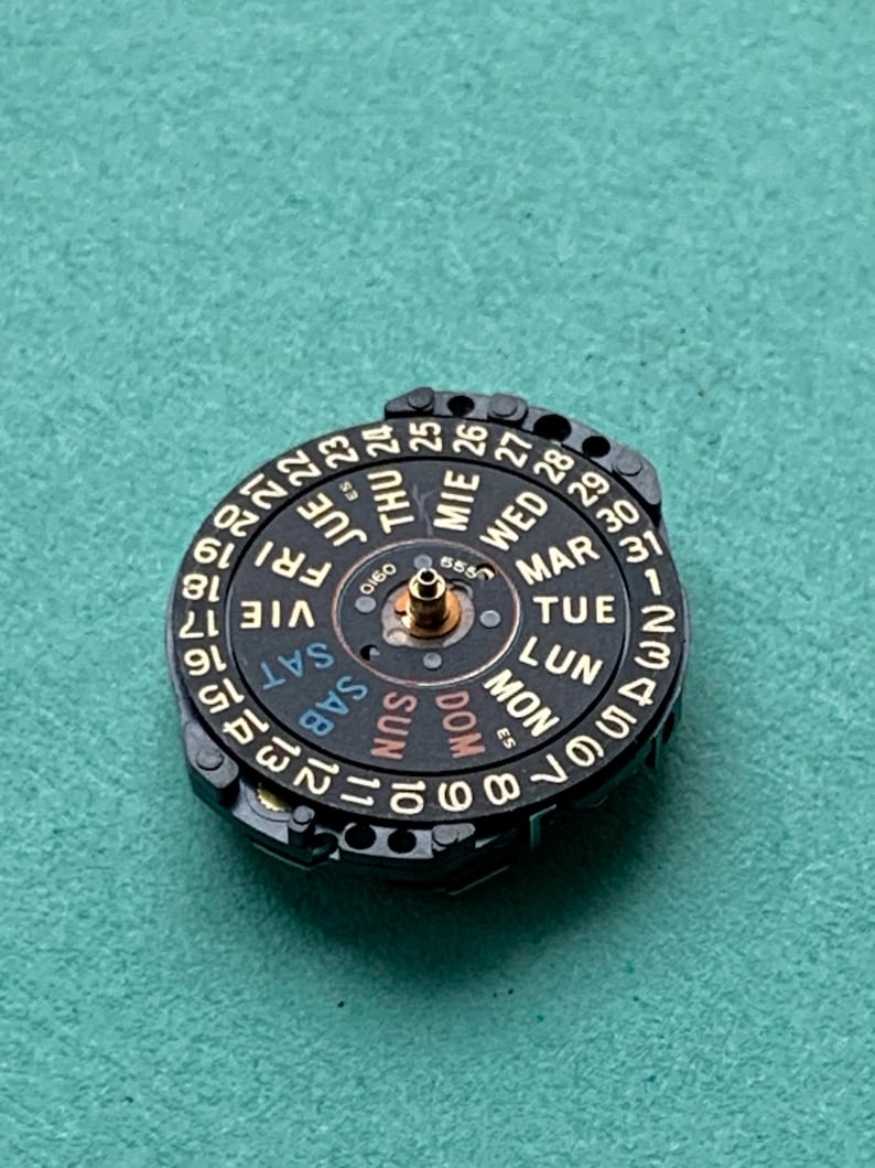 May include: A black watch calendar wheel with gold-colored text and numbers. The wheel displays the days of the week in multiple languages and the dates from 1 to 31. The wheel is set against a teal background.