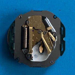 May include: A close-up of a watch movement with gold-coloured gears and components. The watch movement has the text "CITIZEN WATCH CO. JAPAN" and "ONE 1 JEWEL UNADJUSTED". The watch movement is on a blue surface.