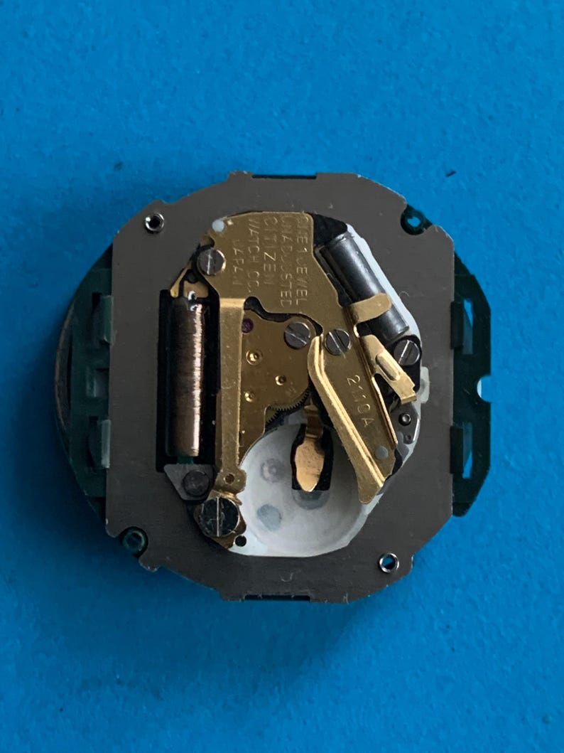 May include: A close-up of a watch movement with gold-coloured gears and components. The watch movement has the text "CITIZEN WATCH CO. JAPAN" and "ONE 1 JEWEL UNADJUSTED". The watch movement is on a blue surface.