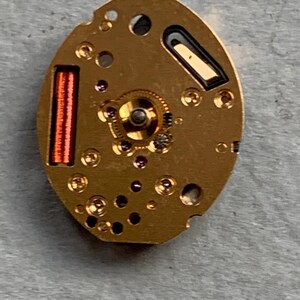 May include: A close-up of a gold-coloured watch movement. The oval mechanism has a central gear, small jewels, and a red coil. Black and dark grey details are also visible.