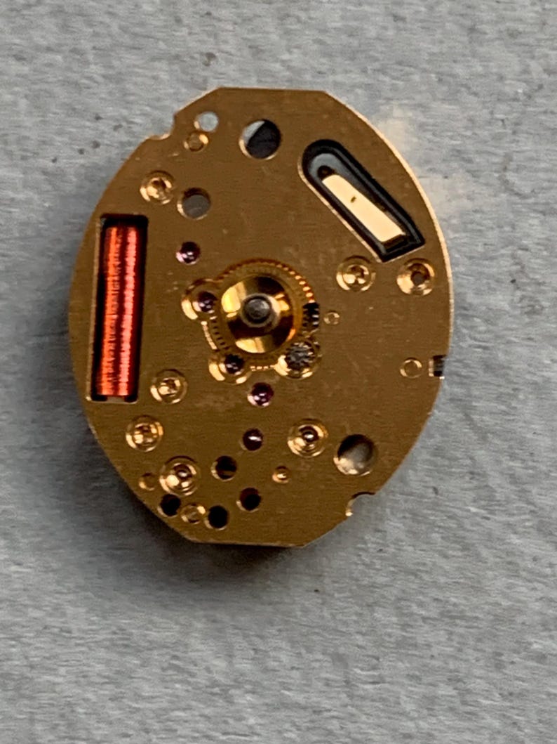 May include: A close-up of a gold-coloured watch movement. The oval mechanism has a central gear, small jewels, and a red coil. Black and dark grey details are also visible.