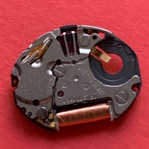 May include: A close-up of a watch movement with silver-colored metal components, including gears, screws, and a copper-colored coil. The watch mechanism has the text "ORIENT JAPAN" and "ONE (1) JEWEL ADJUSTED" printed on it. The background is a solid red.