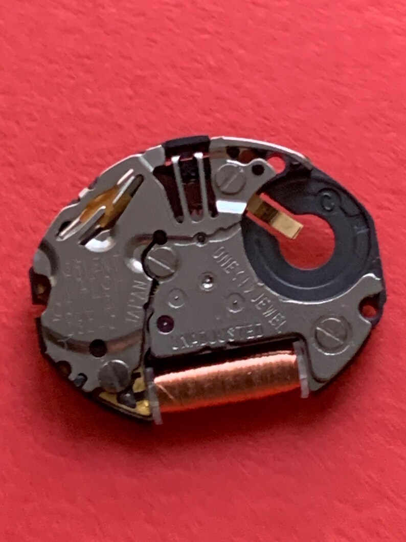 May include: A close-up of a watch movement with silver-colored metal components, including gears, screws, and a copper-colored coil. The watch mechanism has the text "ORIENT JAPAN" and "ONE (1) JEWEL ADJUSTED" printed on it. The background is a solid red.