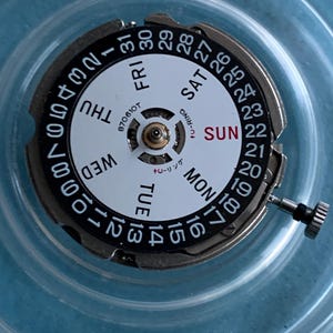 May include: A detailed view of a watch movement, featuring a white dial with black numerals and the days of the week. The date is also displayed. A small crown is present. The text "870610T" is visible.