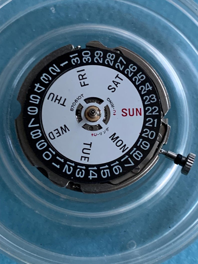 May include: A detailed view of a watch movement, featuring a white dial with black numerals and the days of the week. The date is also displayed. A small crown is present. The text "870610T" is visible.