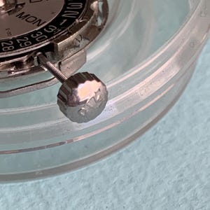 May include: Close-up of a watch movement with a silver-colored dial and a date display. The dial shows the days of the week and the date. A silver crown is visible. The watch parts are inside a clear plastic container.