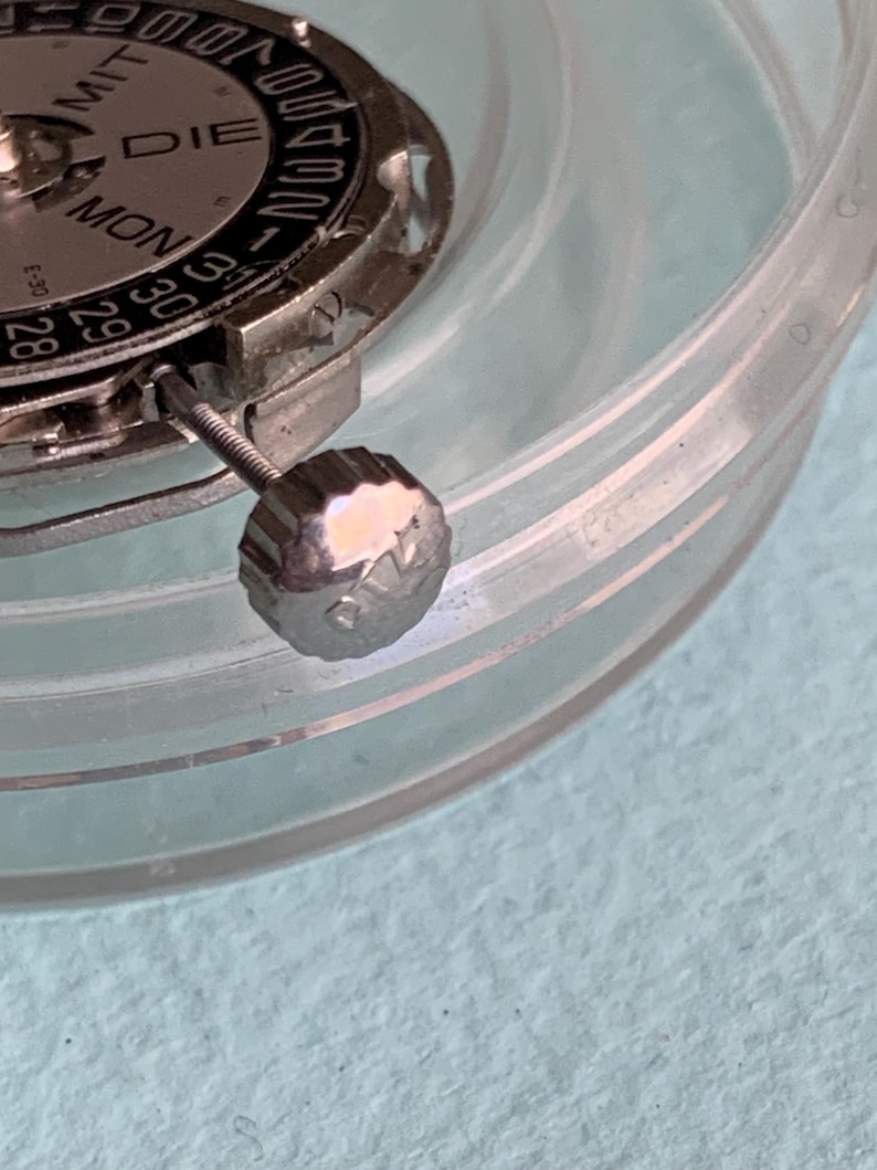 May include: Close-up of a watch movement with a silver-colored dial and a date display. The dial shows the days of the week and the date. A silver crown is visible. The watch parts are inside a clear plastic container.