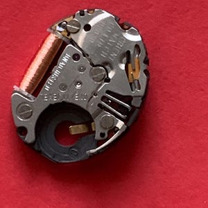May include: Close-up of a watch movement with silver and gold components. A copper-colored coil is visible, along with small screws and gears. The watch mechanism is set against a red background.