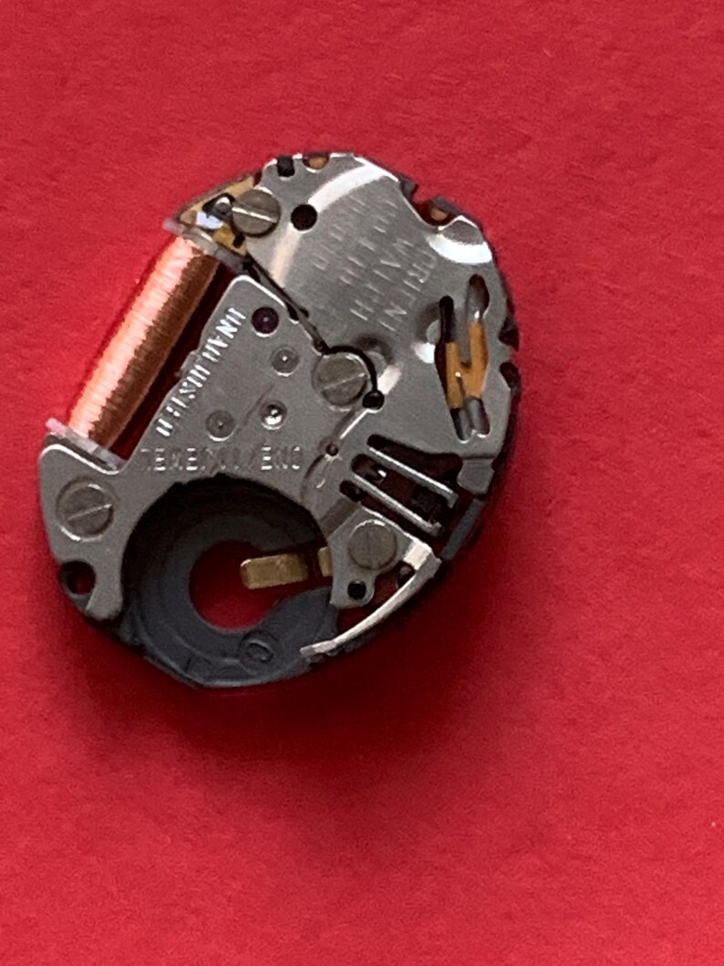 May include: Close-up of a watch movement with silver and gold components. A copper-colored coil is visible, along with small screws and gears. The watch mechanism is set against a red background.