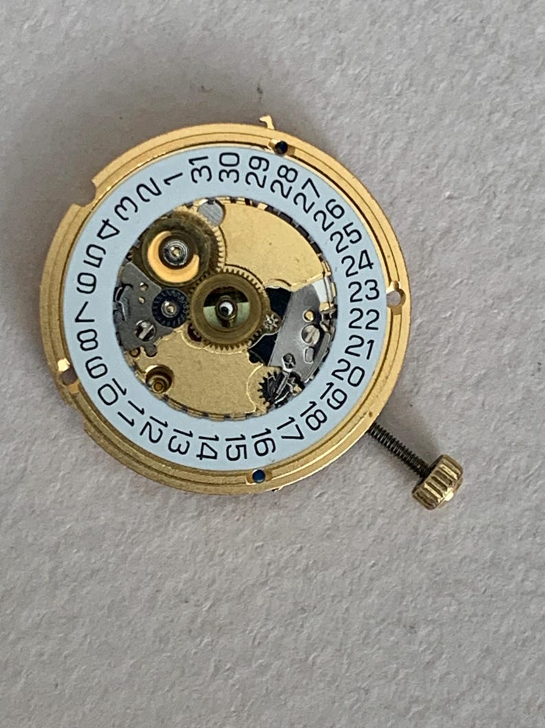 May include: A gold-toned watch movement with intricate gears and a light blue ring displaying the days of the month. A small screw extends from the side. The watch mechanism is a detailed example of horological engineering.