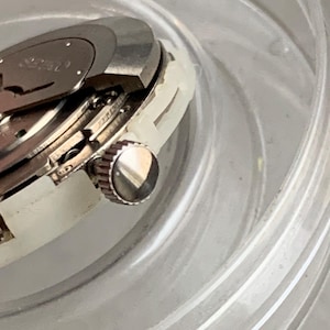May include: Close-up of a silver watch mechanism with a crown and internal gears. The watch is partially disassembled, revealing intricate details of its inner workings. The watch is set in a clear, plastic container.