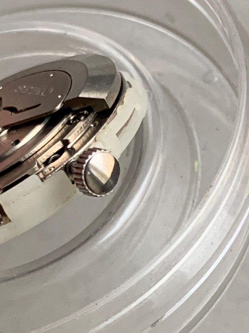 May include: Close-up of a silver watch mechanism with a crown and internal gears. The watch is partially disassembled, revealing intricate details of its inner workings. The watch is set in a clear, plastic container.