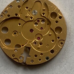 May include: Close-up of a gold-colored watch movement with intricate gears, holes, and a visible serial number 2571. The movement features small red jewels and various mechanical components, showcasing the internal workings of a timepiece.