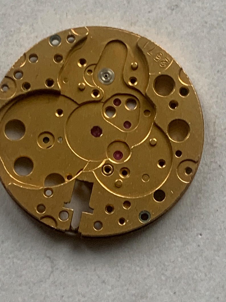 May include: Close-up of a gold-colored watch movement with intricate gears, holes, and a visible serial number 2571. The movement features small red jewels and various mechanical components, showcasing the internal workings of a timepiece.