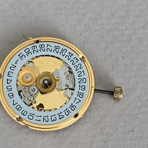 May include: A gold-toned watch movement with a light blue date dial and intricate gears. The dial features black numerals from 1 to 31. A small screw and crown are attached to the side. The watch mechanism is designed for repair or restoration.