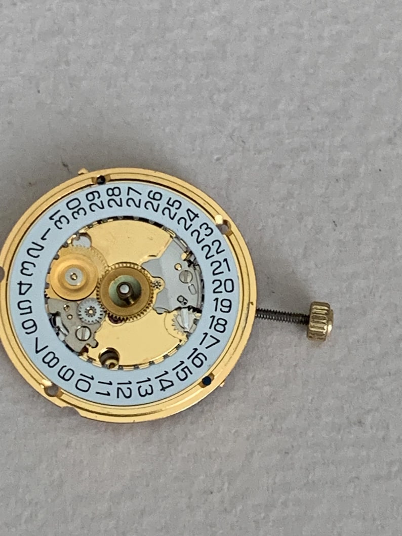 May include: A gold-toned watch movement with a light blue date dial and intricate gears. The dial features black numerals from 1 to 31. A small screw and crown are attached to the side. The watch mechanism is designed for repair or restoration.