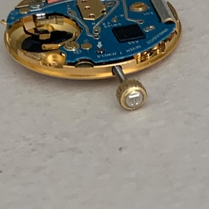 May include: Close-up of a watch movement with a gold-colored frame and a blue circuit board. The watch mechanism has a small screw and the words "SWISS V8" and "SEVEN 7 JEWELS" are visible.
