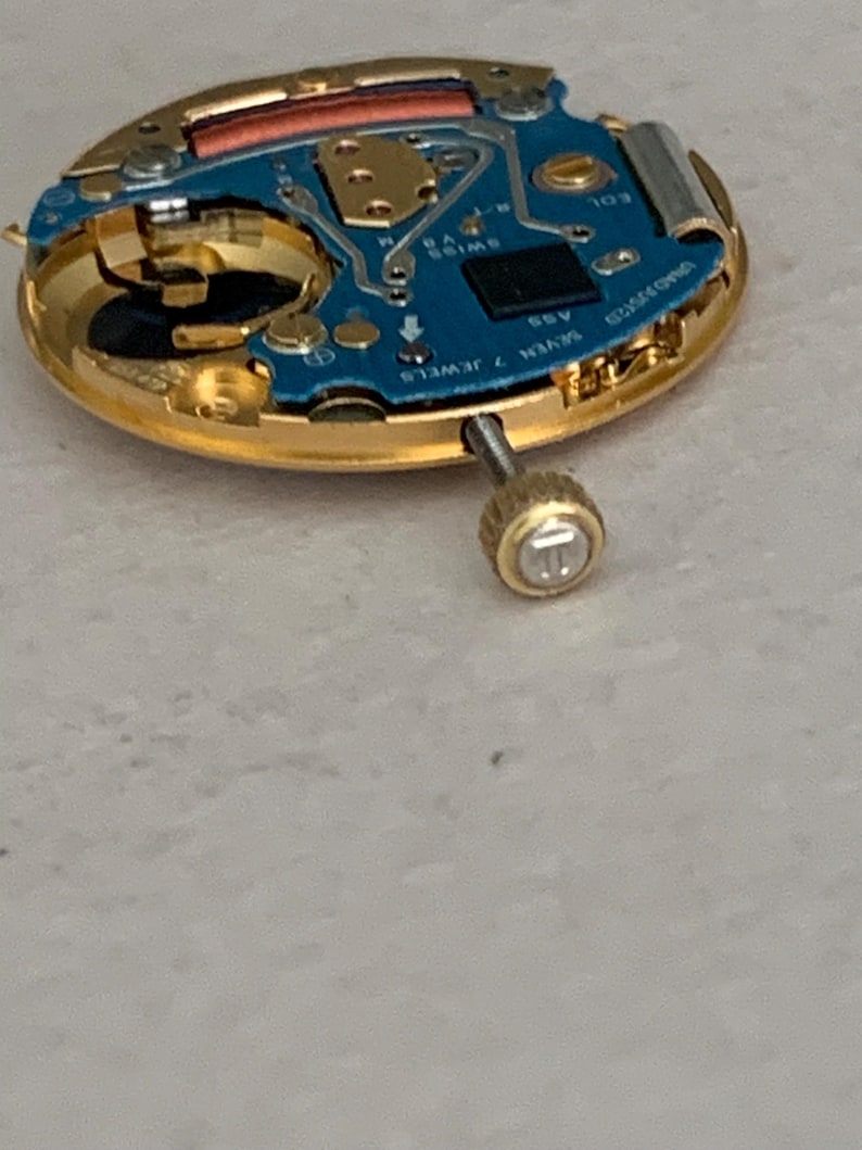 May include: Close-up of a watch movement with a gold-colored frame and a blue circuit board. The watch mechanism has a small screw and the words "SWISS V8" and "SEVEN 7 JEWELS" are visible.