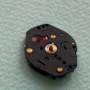 May include: Close-up of a black watch movement with gold-coloured accents. The movement features a small, central gear and several other components. The background is a light green colour.