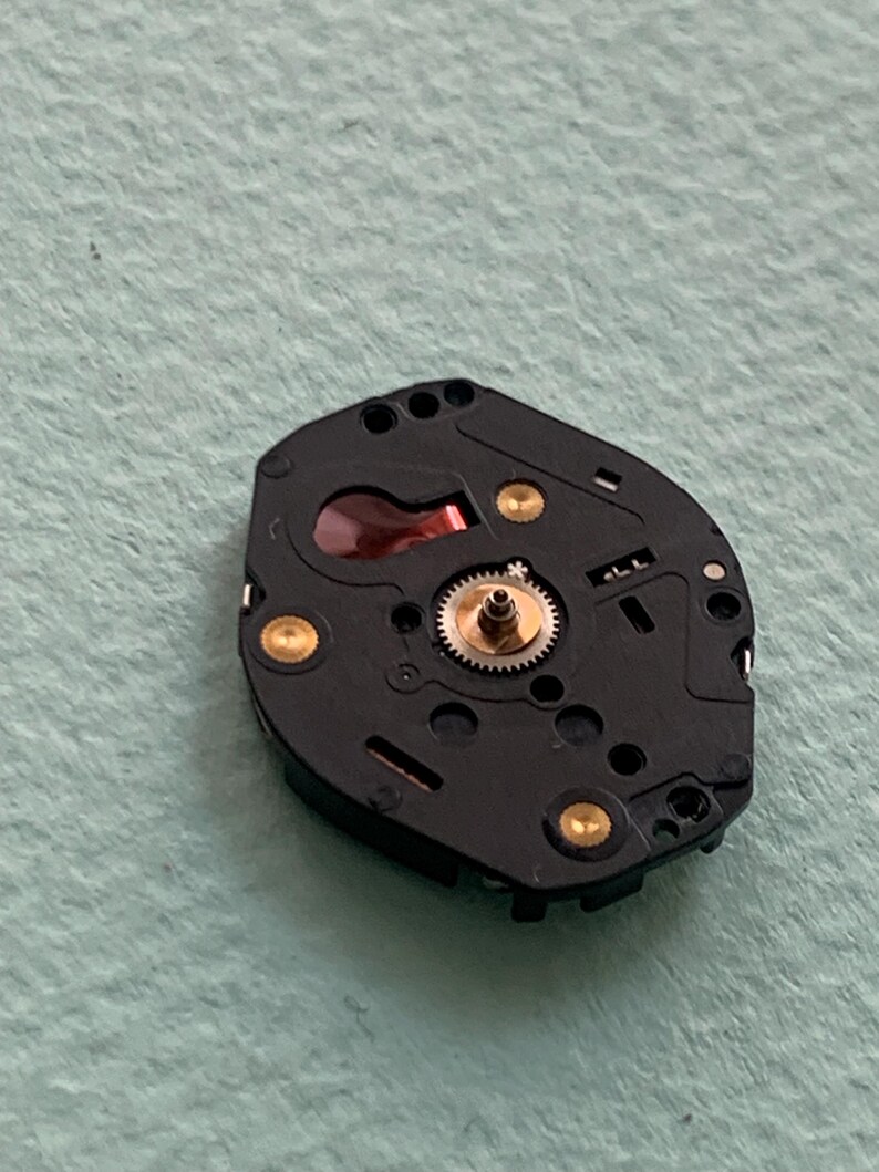 May include: Close-up of a black watch movement with gold-coloured accents. The movement features a small, central gear and several other components. The background is a light green colour.