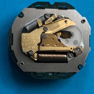 May include: A watch movement featuring gold-coloured gears and a silver-coloured base. The movement is inscribed with "ONE JEWEL UNADJUSTED CITIZEN JAPAN". This is a mechanical watch component.