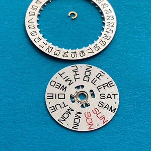 May include: Watch parts, including a white date wheel with black numbers and a day wheel with days of the week in black and red. A small gold-colored component is also present. The parts are on a blue surface.
