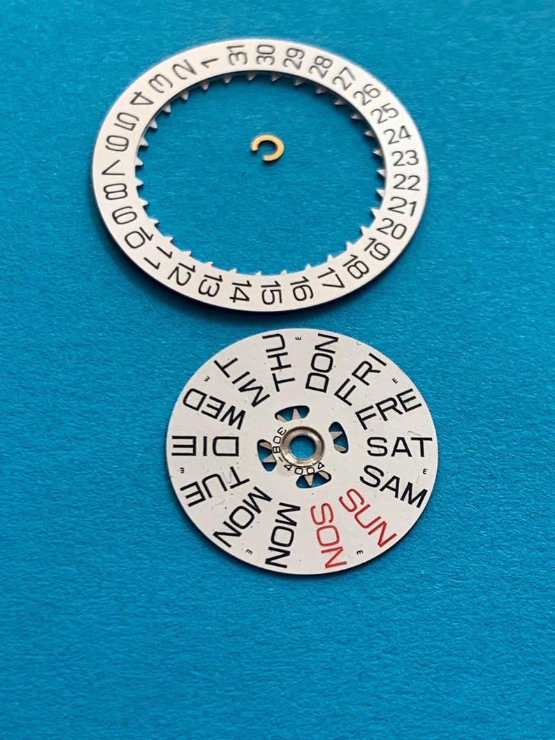 May include: Watch parts, including a white date wheel with black numbers and a day wheel with days of the week in black and red. A small gold-colored component is also present. The parts are on a blue surface.