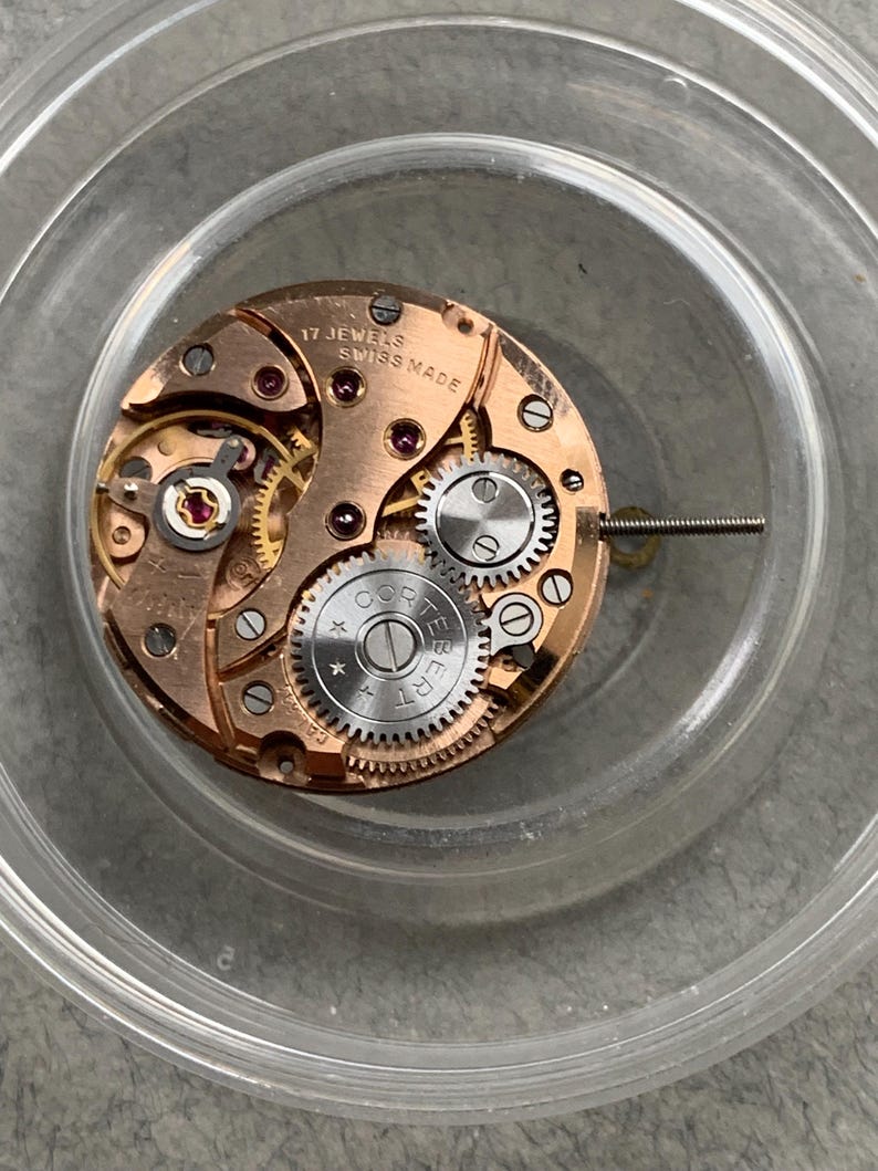 May include: A close-up of a watch movement with visible gears, screws, and jewels. The watch mechanism is copper-colored with the text "17 JEWELS SWISS MADE" and the brand name "CORTEBERT" visible. The watch movement is inside a clear plastic container.