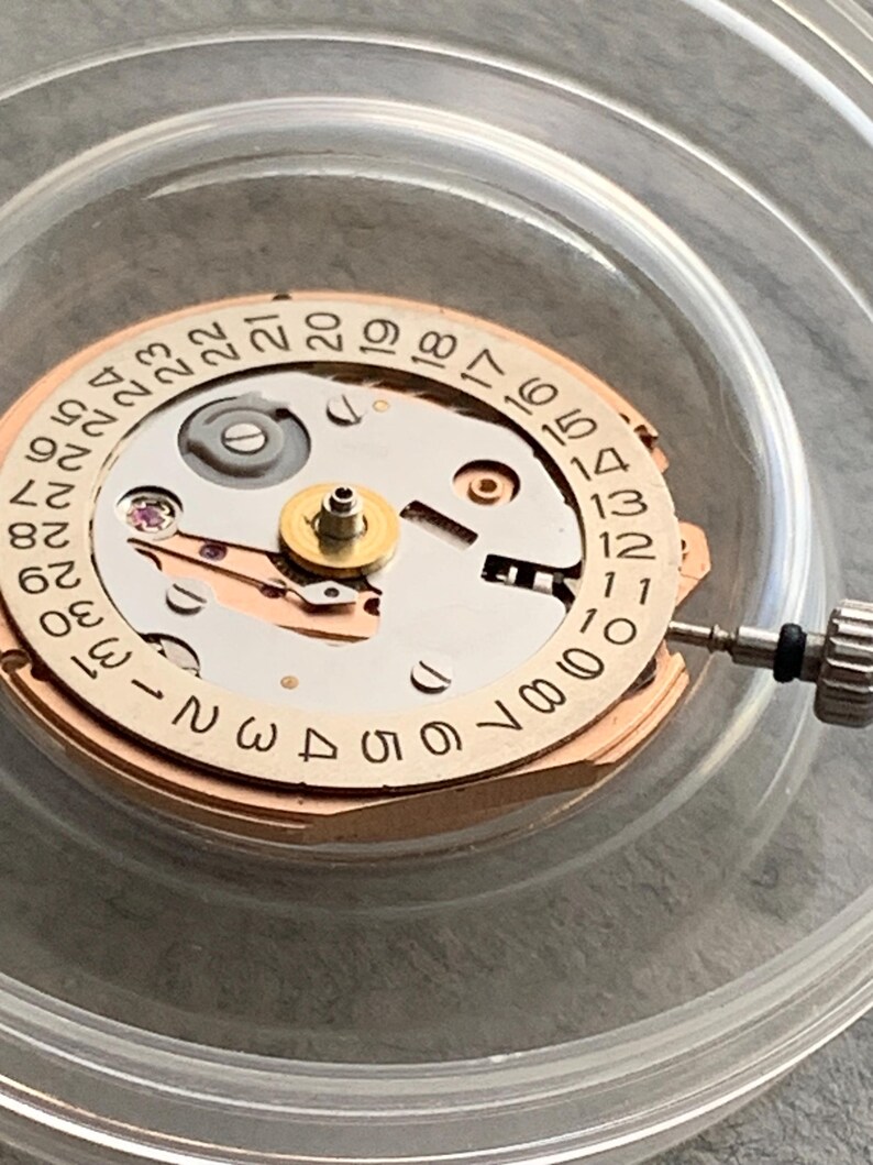 Puede incluir: Primer plano de un movimiento de reloj con una esfera de fecha circular. La esfera es beige con n&uacute;meros negros y un centro plateado. Una peque&ntilde;a herramienta est&aacute; colocada en el lado derecho. El mecanismo del reloj est&aacute; dentro de un recipiente de pl&aacute;stico transparente.
