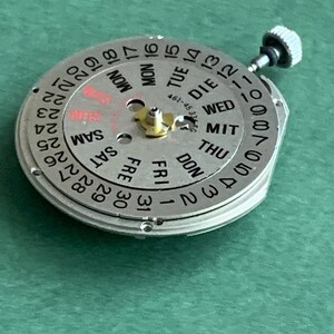 May include: A silver watch movement with a calendar dial showing days and dates. The dial features black numerals and text, with 'SUN' highlighted in red. A small crown is visible. The movement is approximately 3 cm in diameter.