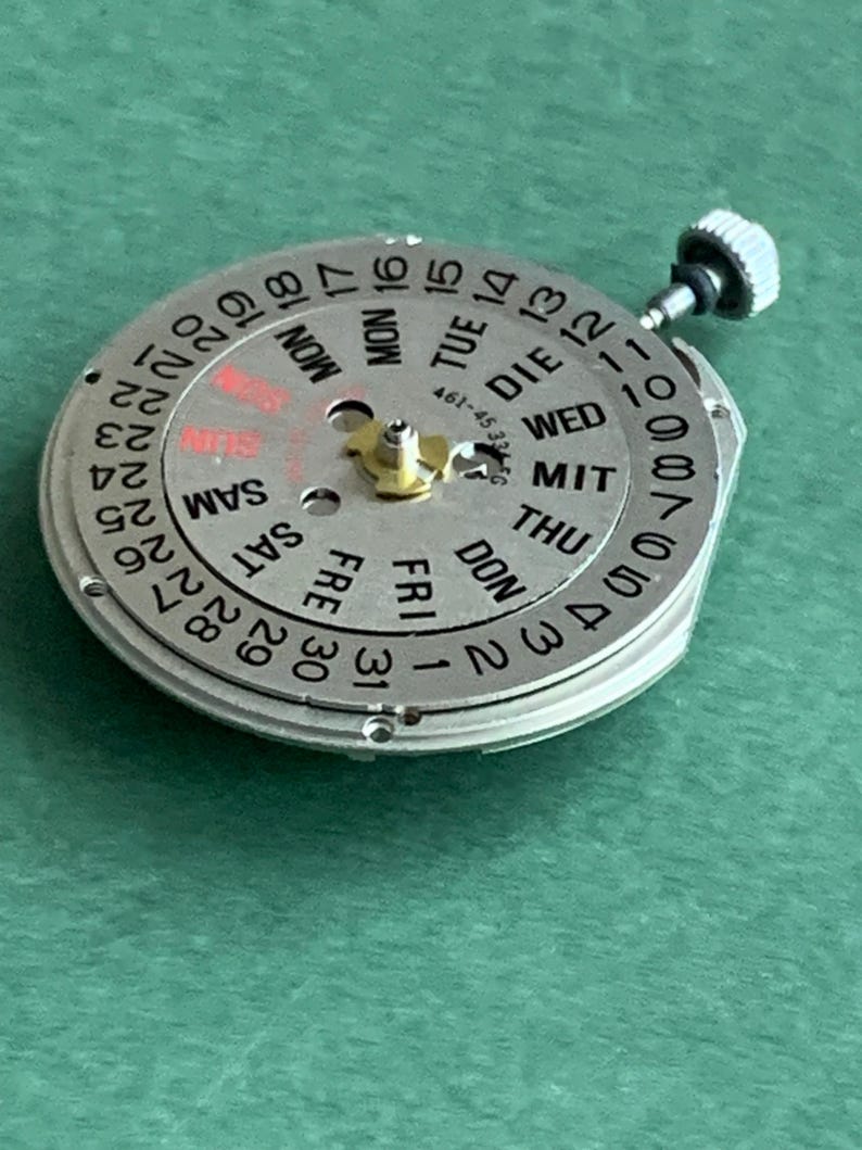 May include: A silver watch movement with a calendar dial showing days and dates. The dial features black numerals and text, with 'SUN' highlighted in red. A small crown is visible. The movement is approximately 3 cm in diameter.