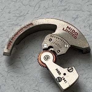 Puede incluir: Un movimiento de reloj plateado con el texto "Mido Limited" y "Seventeen Jewels" impreso en él. El movimiento también tiene el texto "Made in Switzerland" y "Mod. 1941" impreso en él.