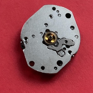 May include: A close-up of a watch movement component. The silver-colored metal piece has several small holes and a central gear with gold-colored teeth. The background is a solid red.