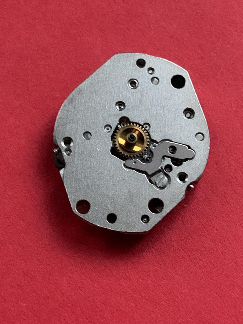 May include: A close-up of a watch movement component. The silver-colored metal piece has several small holes and a central gear with gold-colored teeth. The background is a solid red.