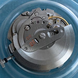 May include: Close-up of a disassembled watch movement. The intricate mechanism features silver-coloured gears, screws, and plates. The watch movement is set against a light blue background, showcasing the detailed craftsmanship of the timepiece.
