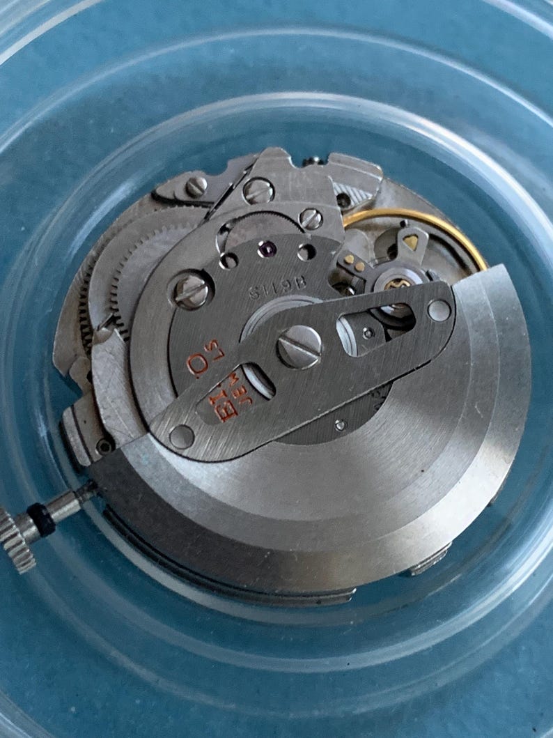 May include: Close-up of a disassembled watch movement. The intricate mechanism features silver-coloured gears, screws, and plates. The watch movement is set against a light blue background, showcasing the detailed craftsmanship of the timepiece.