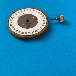 May include: A watch movement with a brown outer ring and a white inner ring displaying numbers 1-31. The center has a small, metallic mechanism. A gold-colored crown and stem extend from the side. The background is a solid blue.