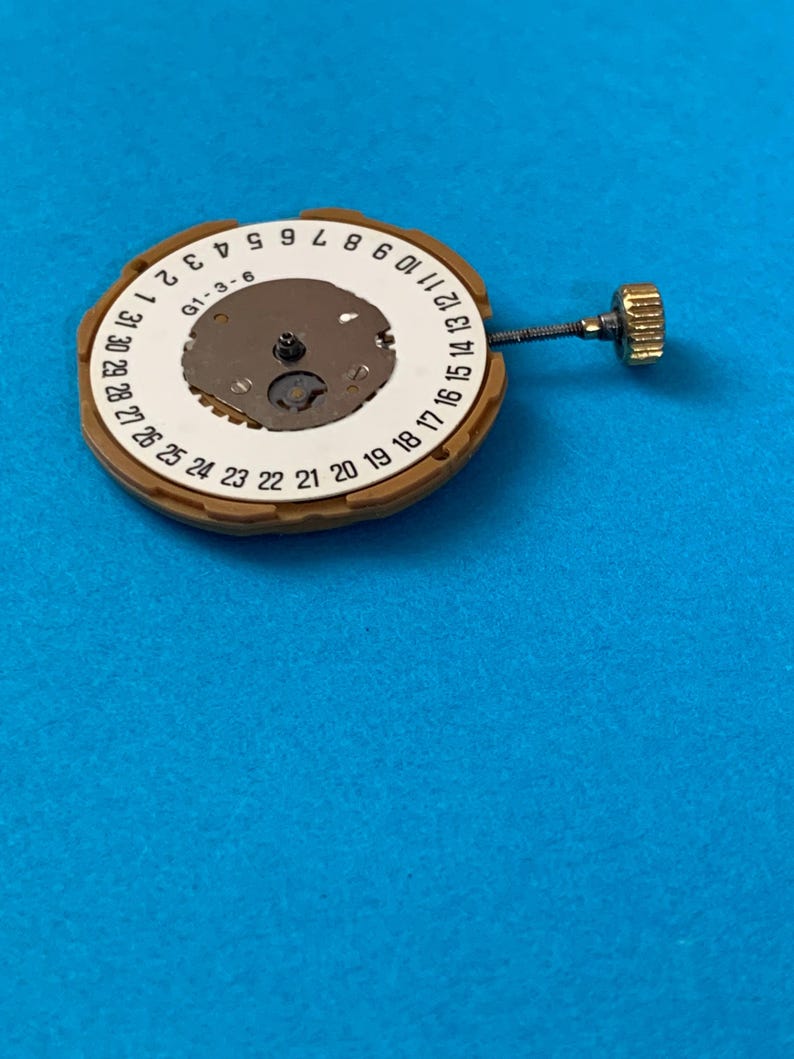 May include: A watch movement with a brown outer ring and a white inner ring displaying numbers 1-31. The center has a small, metallic mechanism. A gold-colored crown and stem extend from the side. The background is a solid blue.