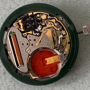 May include: Close-up of a Citizen watch movement with intricate gold-colored gears and components. The movement is housed in a dark green circular case. The watch mechanism includes a red battery and the text "JAPAN FIVE 5 ADJUSTED JEWELS".