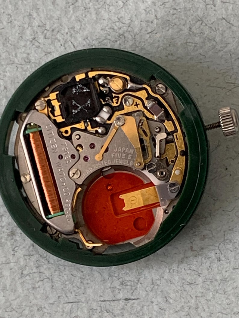 May include: Close-up of a Citizen watch movement with intricate gold-colored gears and components. The movement is housed in a dark green circular case. The watch mechanism includes a red battery and the text "JAPAN FIVE 5 ADJUSTED JEWELS".