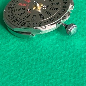 May include: Close-up of a silver watch movement with a day and date display. The dial features the days of the week and numbers for the date. The background is a solid green color.