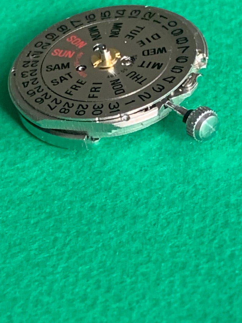 May include: Close-up of a silver watch movement with a day and date display. The dial features the days of the week and numbers for the date. The background is a solid green color.