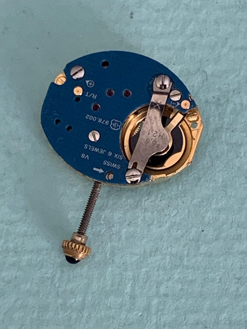 May include: A disassembled watch movement with a blue circuit board and gold-colored components. The board has text including "SWISS SIX 6 JEWELS" and "976.002". A screw and a small black and gold crown are also visible.