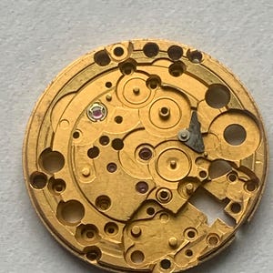 May include: Close-up of a gold-colored watch movement with intricate gears, jewels, and components. The circular mechanism features numerous holes and small details, showcasing the internal workings of a timepiece. The image highlights the precision and complexity of the watch's internal structure.